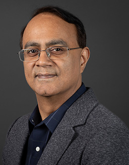 Male person wearing glasses in a dark shirt and light jacket on a dark background