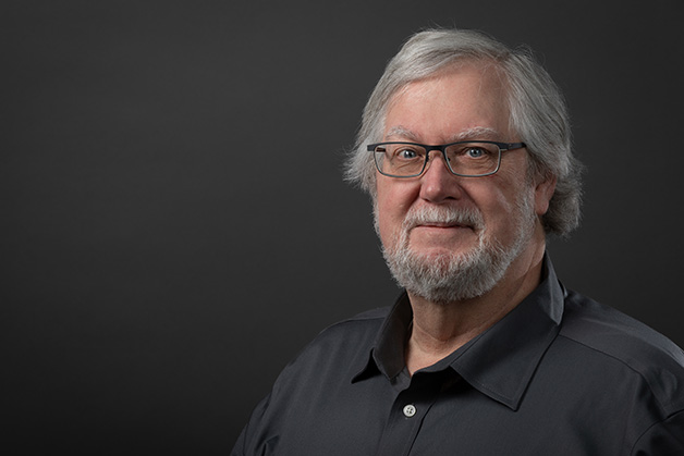 Male person with glasses and a beard in a gray shirt on a dark background