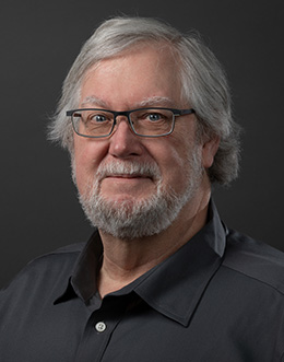 Male person with glasses and a beard in a gray shirt on a dark background