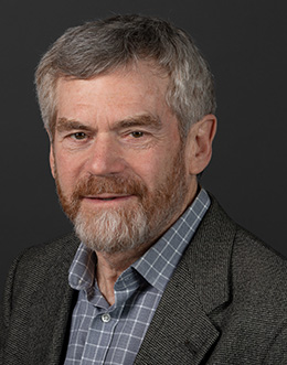 Male person with a beard in a patterned shirt and a dark jacket on a dark background